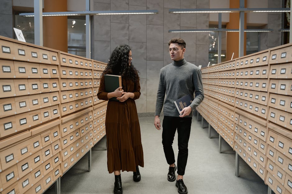 Two students walk through a library archive, each holding books and conversing.